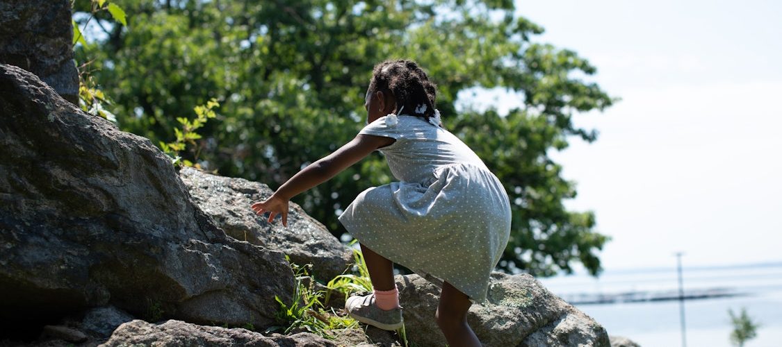 An African American girl climbing a rock outside in a dress.