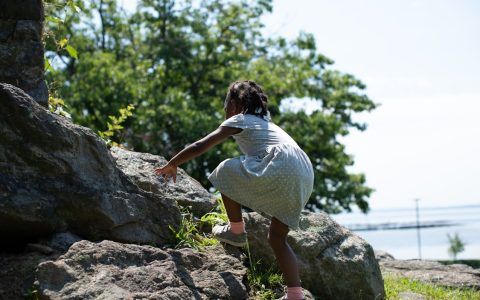 An African American girl climbing a rock outside in a dress.